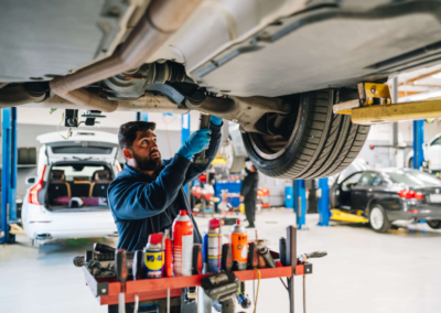 Bearded mechanic in a blue work uniform and blue gloves reaching up to service the undercarriage of a car on a lift, with a busy multi-bay auto shop in the background and a red service cart stocked with WD-40 and lubricants in the foreground