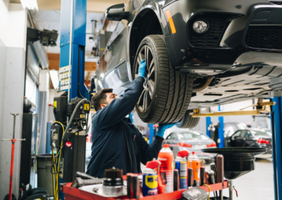 Mechanic in a dark blue uniform and blue gloves inspecting the undercarriage of a black SUV raised on a hydraulic lift, with a red service cart holding WD-40 and other automotive fluids in the foreground and multiple bays visible behind